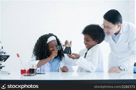 Little African kids studying chemistry and doing chemical science experiment in laboratory at school. Dark skinned girl using magnifying glass to look at liquid in test bottle with Asian man teacher