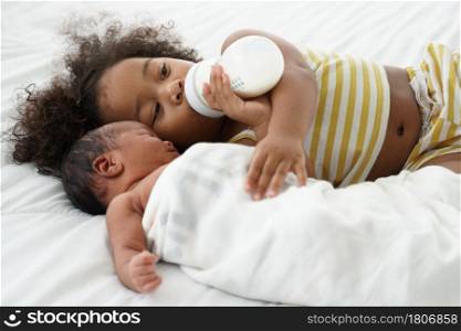 Little African older sister sucking milk from bottle, hugging and take good care of her newborn baby brother lying on white bed wrap with blanket at bedroom at home. Relationship of small siblings.