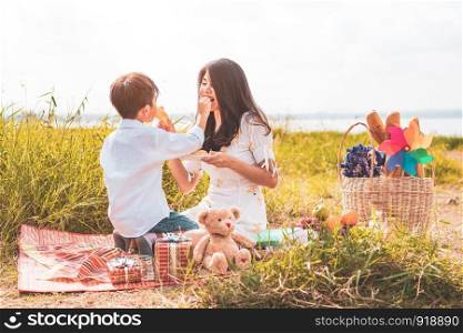 Little Asian boy his mom feeding snack each other in meadow when doing picnic. Mother and son playing together. Celebrating in Mother day and appreciating concept. Summer people and lifestyle theme.