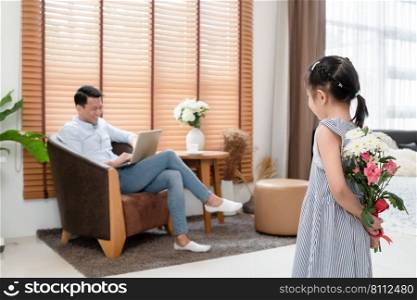 Little Asian cute daughter holding hiding bunch of flowers behind her back for surprise her father who is working with laptop at living room at modern home. Happy father"s day.
