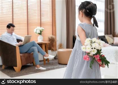 Little Asian cute daughter holding hiding bunch of flowers behind her back for surprise her father who is working with laptop at living room at modern home. Happy father’s day.