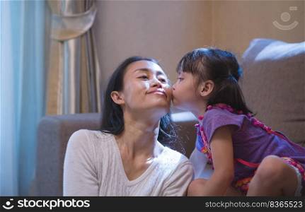 Little Asian girl on couch kissing her mother"s cheek in living room at home