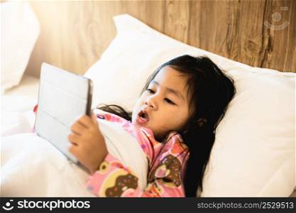 Little Asian girl playing digital tablet on bed addicted game and cartoon, Kid watching videos and cartoons online in bedroom at home, Fun online concept