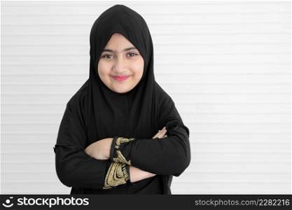 Little Asian Pakistani Muslim girl wearing black hijab with beautiful eyes is smiling and standing with arms crossed. White background