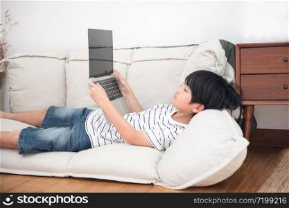 Little asian school boy in white shirt laying in his bed play on laptop. Social network addiction concept. Child pc dependence
