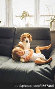Little baby girl with beagle dog sitting on the sofa at home. Pets at home concept background.. Little baby girl with beagle dog sitting on the sofa at home.