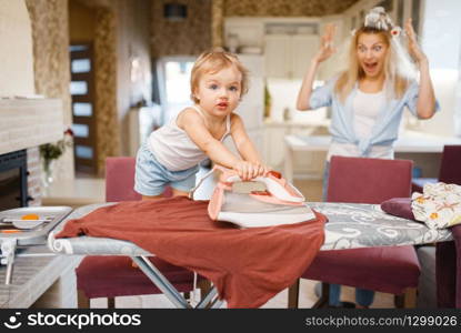 Little baby irons clothes on the kitchen, housewife in horror. Kid doing housework at home. Young child clean the house