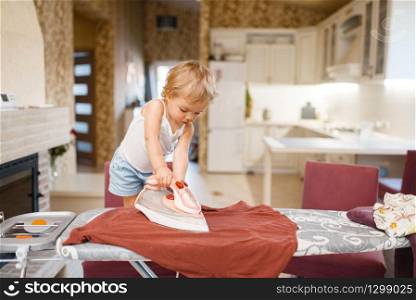 Little baby irons clothes on the kitchen. Kid doing housework at home. Young housewife clean the house