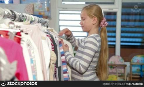 Little blond girl shopping for clothes in a clothing store, looking sweater. Side view