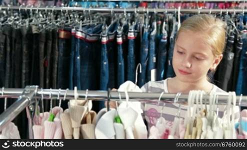 Little blond girl shopping for clothes in a clothing store, trying on sweater. Front view