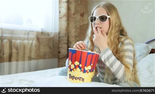 Little blonde girl eating popcorn and watching 3D movie at home