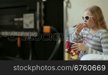 Little blonde girl eating popcorn and watching 3D movie at home, dolly shot