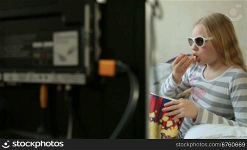 Little blonde girl eating popcorn and watching 3D movie at home, dolly shot