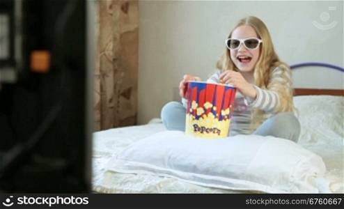 Little blonde girl eating popcorn and watching 3D movie at home, screaming and laughing
