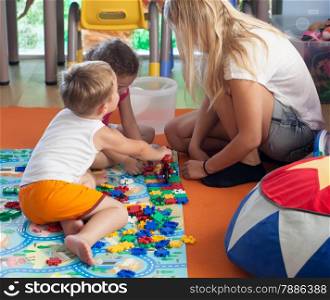Little boy and girl playing with schoolmaster in game room or nursery. Girl doing puzzle, boy playing with toy bike