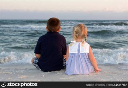 Little boy and girl sit at the beach and watch the waves roll in at sunset.