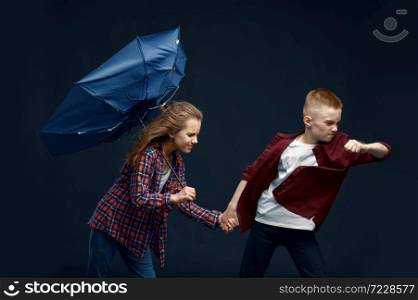 Little boy and girl with umbrella against powerful airflow in studio, wind effect, windy. Children with developing hairs, kids isolated on dark background, child emotion. Little boy and girl with umbrella against airflow