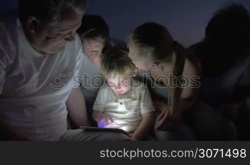 Little boy and his family are sitting together with tablet PC at night. They are talking and boy is looking for something in the gadget.