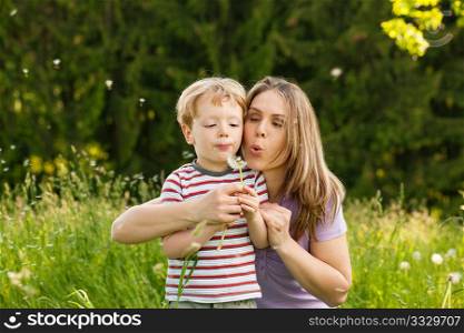 Little boy and his mother blowing dandelion seed for a wish on a meadow outdoors in summer