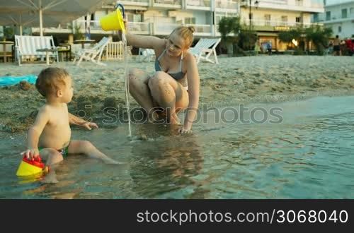 Little boy and his mother playing on the beach in the shallow water in front of an apartment block or resort hotel as they enjoy a summer evening