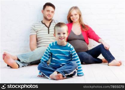 Little boy and his parents sitting on the floor near the wall. Mother is pregnant.