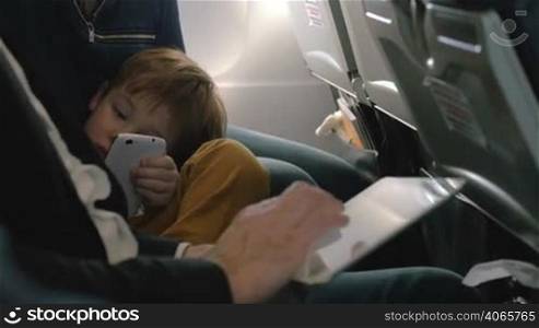 Little boy bored with flight using smartphone lying on mothers lap and his father working with tablet computer