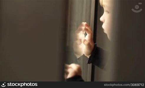 Little boy coming close to the window and looking out of with his own reflection there