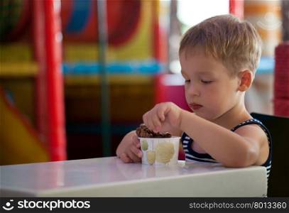 Little boy having tasty chocolate ice cream sitting at the table in game room