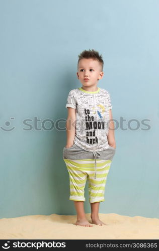 little boy in a shirt and shorts standing on the sand on a light blue background. little boy in shorts