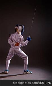 Little boy in a white protective suit fencing mask and with a rapier exercises in fencing on a dark background. boy engaged in fencing