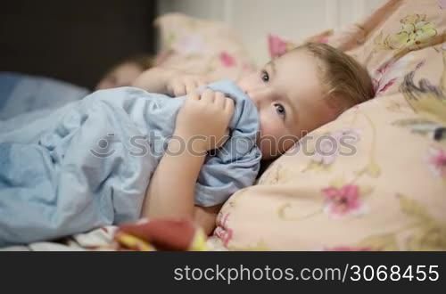Little boy in bed playing with the blanket, putting it into the mouth.