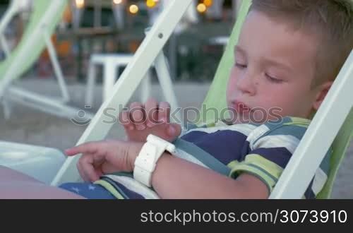 Little boy in chaise-longue at the seaside using smart watch. Modern gadgets for modern generation