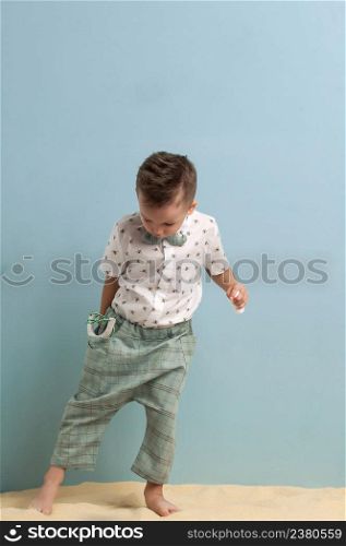 little boy in fashionable clothing and a slingshot in his hands stands on the sand on a light blue background. little boy in a suit