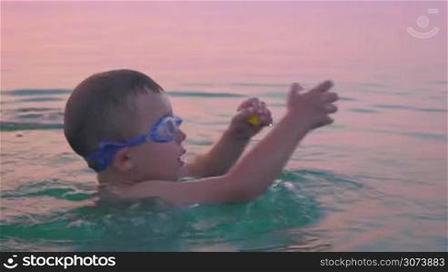 Little boy in goggles turning around himself in sea at sunset