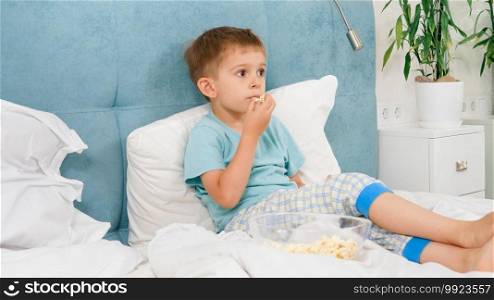 Little boy in pajamas lying in bed and eating popcorn while watching TV at morning. Little boy in pajamas lying in bed and eating popcorn while watching TV at morning.