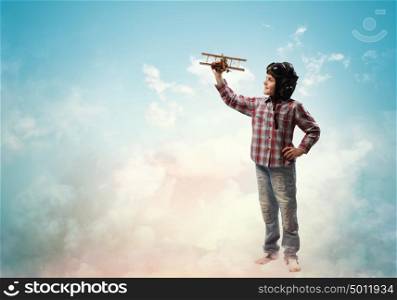 Little boy in pilot&rsquo;s hat. Image of little boy in pilots helmet playing with toy airplane against clouds background