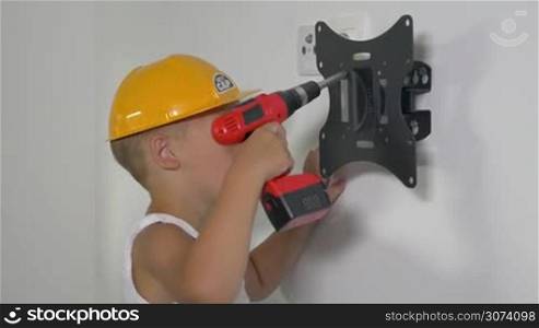 Little boy in safety helmet is fastening wall mount with electric screwdriver.