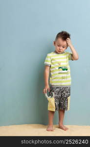 little boy in shorts and a T-shirt with a slingshot in his hands stands on the sand on a light blue background BALIQ - A FISH. little boy in shorts BALIQ - A FISH