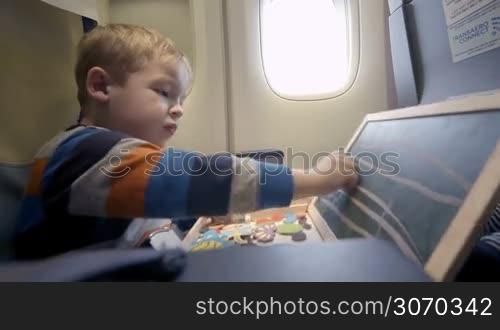 Little boy in the plane drawing on small board with chalk. Entertainment during flight