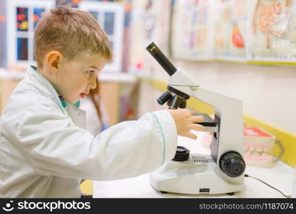 Little boy in uniform sitting at the microscope and playing doctor, playroom. Kid plays medicine worker in imaginary hospital lab, profession learning, childish dream
