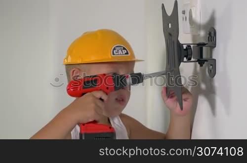 Little boy in yellow hard hat playing a repairer. He using toy screw gun to adjust holder on the wall. Serious small worker