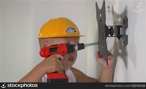 Little boy in yellow hard hat playing a repairer. He using toy screw gun to adjust holder on the wall. Serious small worker