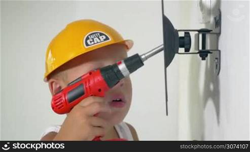 Little boy in yellow safety cap using toy screw gun to fix the holder on the wall. Child pretending to be a repair