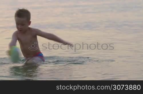 Little boy is playing in the calm sea at sunset, he is holding a toy boat and spinning around.