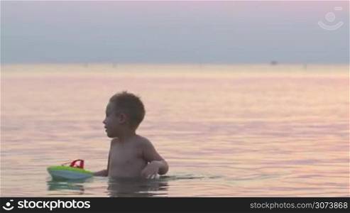 Little boy is playing with toy boat in calm sea or lake at sunset.