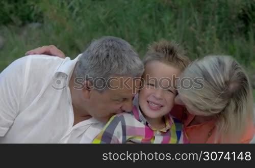 Little boy is sitting between his grandparents, they are hugging and kissing him.