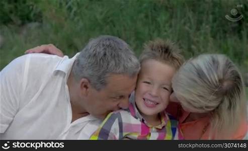 Little boy is sitting between his grandparents, they are hugging and kissing him.