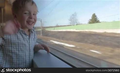 Little boy is standing by the window of moving train and waving hand.