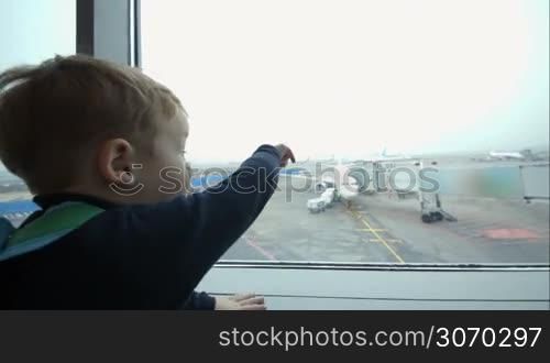 Little boy looking out the window at the airport and pointing at the plane on takeoff strip