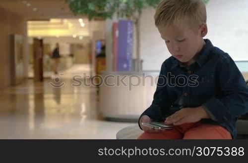 Little boy looking through the photos on smart phone in shopping mall, unidentified people passing by